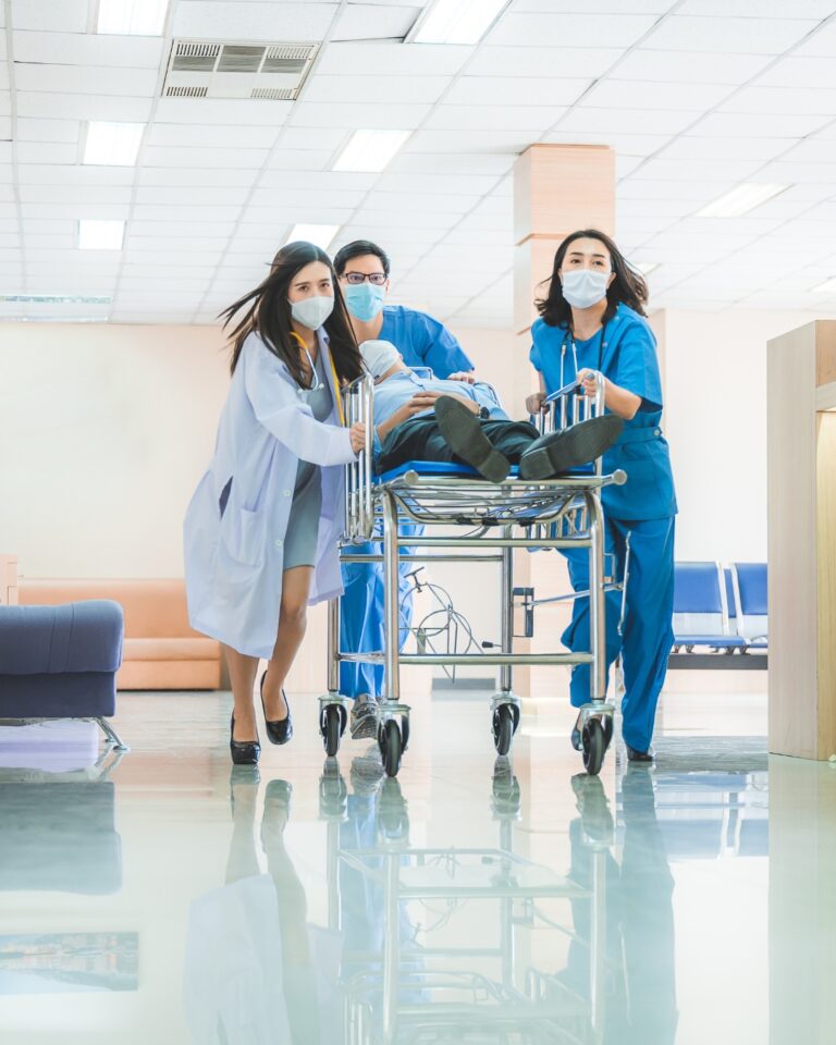 Three healthcare workers, wearing masks, rush a patient on a gurney through a hospital hallway during an emergency.