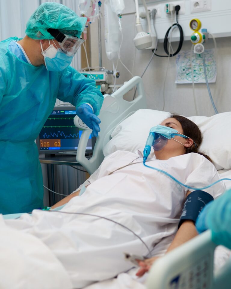 Doctor in PPE checking on patient with oxygen mask in hospital bed.