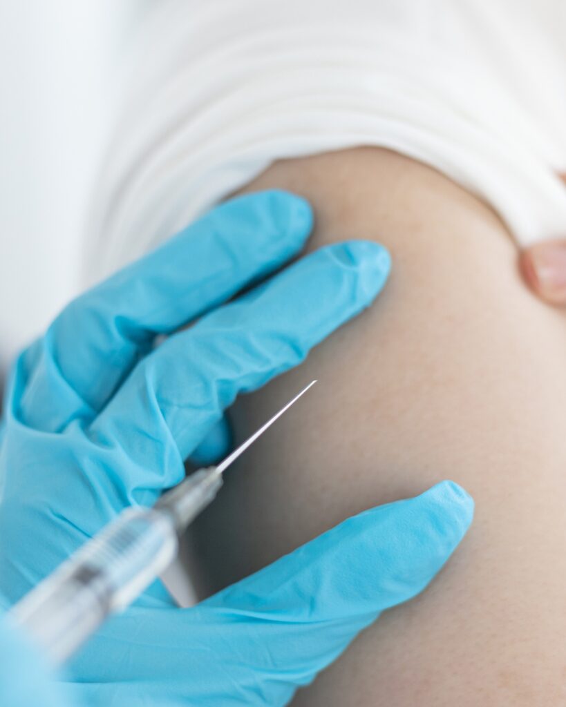 Gloved hand administering injection with syringe and needle into upper arm.